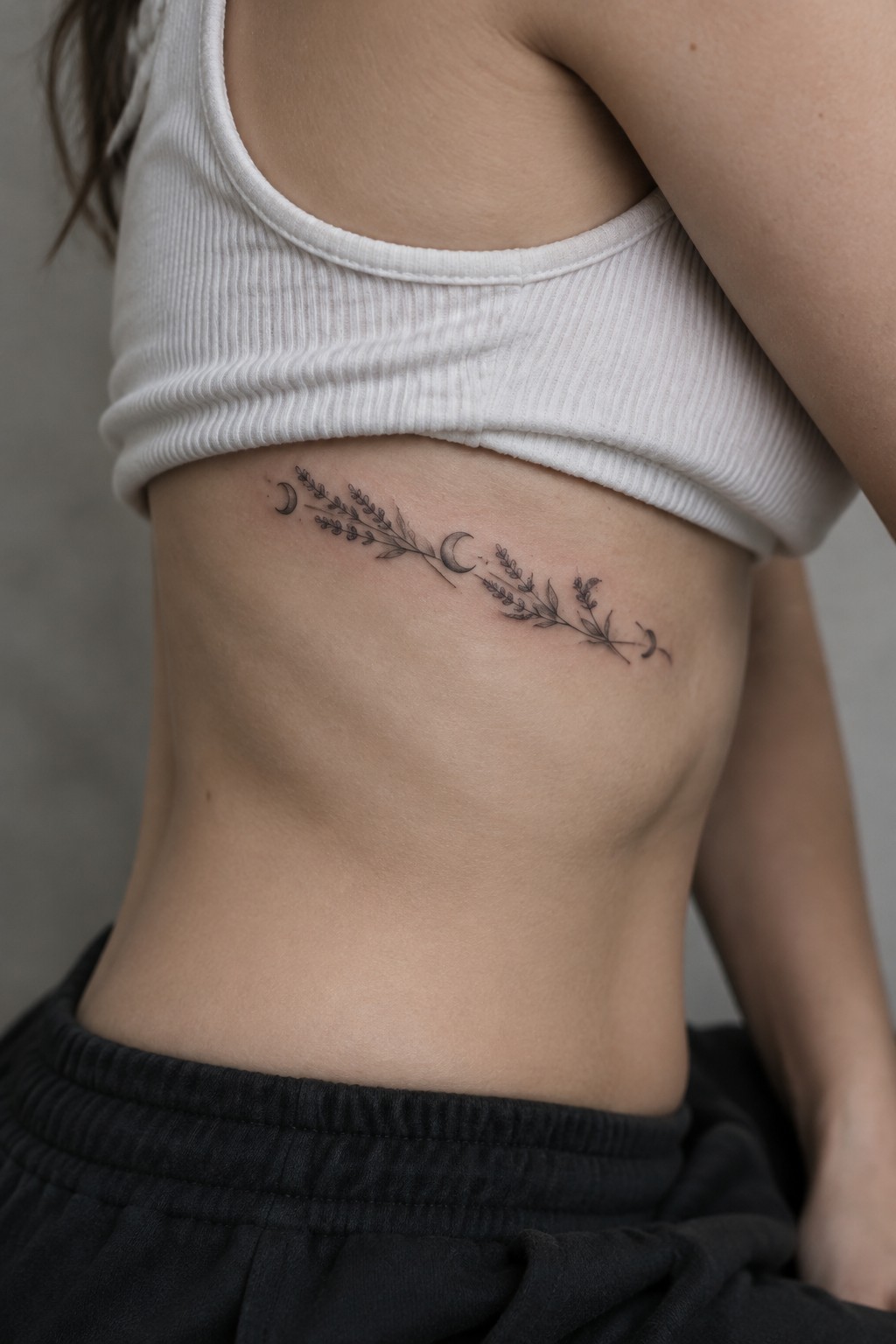 Fine-line leafy branch tattoo with small crescent moons placed along a woman’s ribs beneath a white crop top.