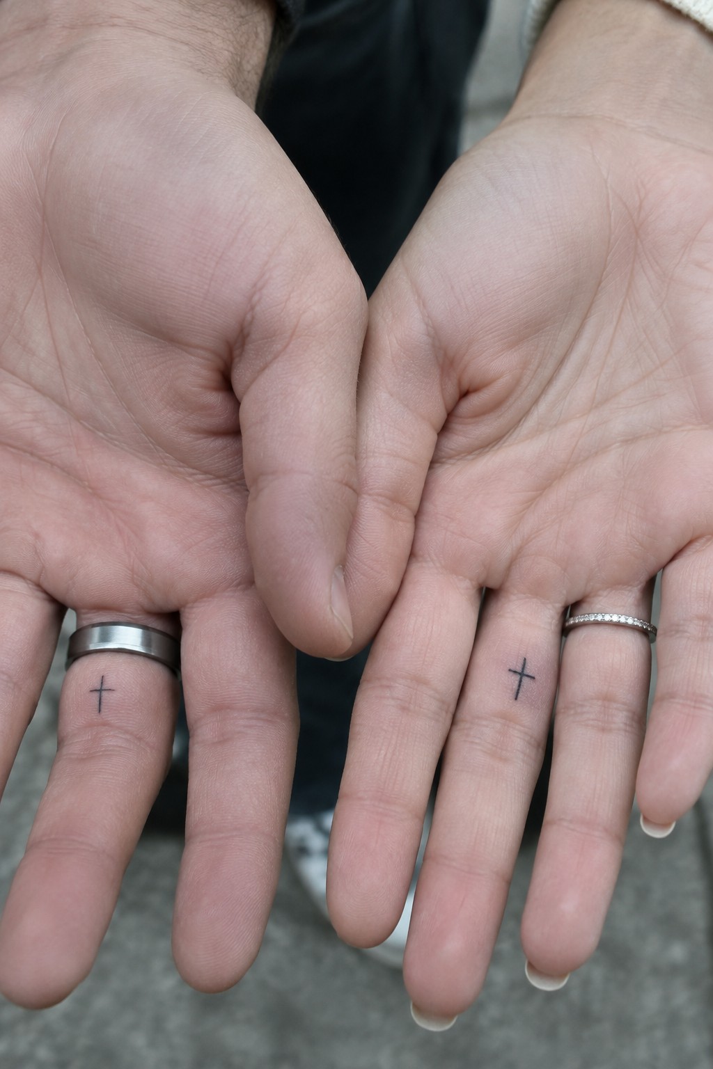 Two hands with matching cross tattoos on ring fingers beside silver and diamond rings