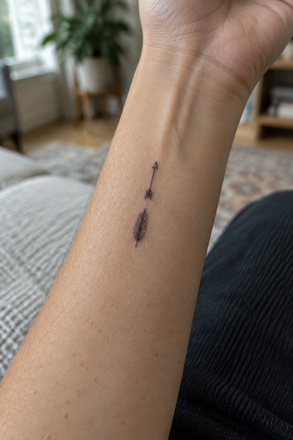 Small black arrow and softly shaded feather tattoo arranged vertically on the inner forearm.