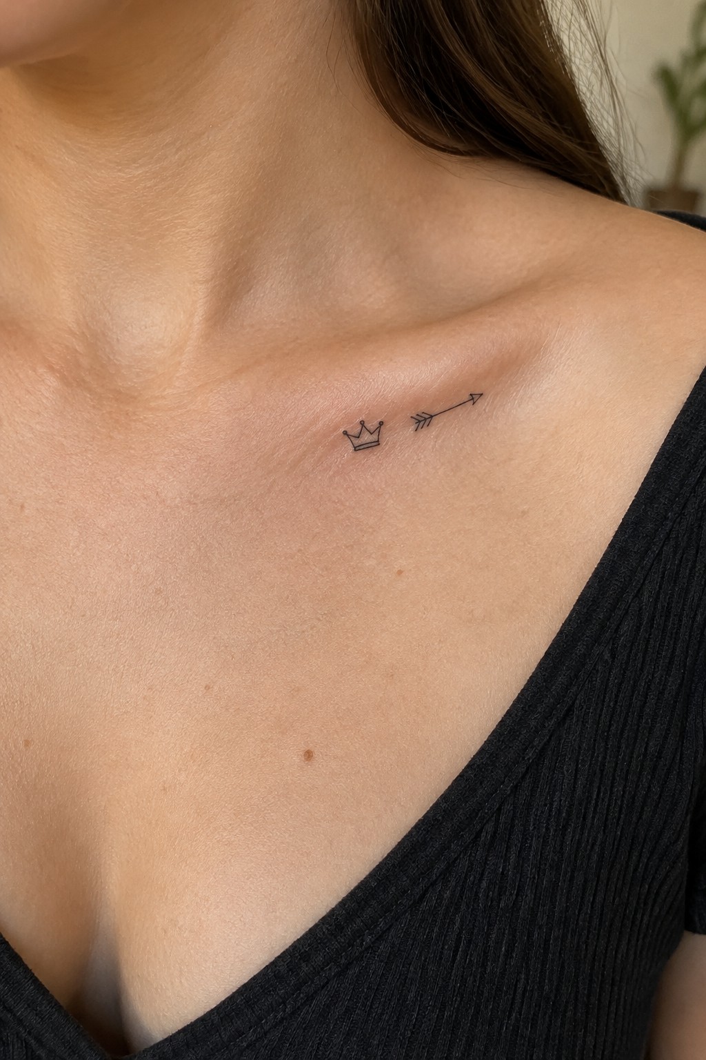 Small black crown and arrow tattoo along a woman’s collarbone.