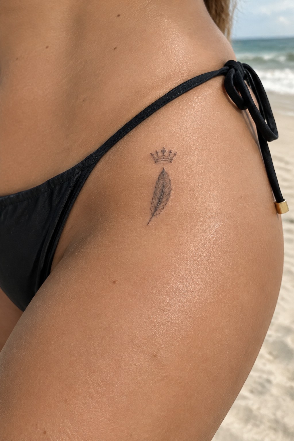 Small black crown and feather tattoo on the outer hip above a black bikini bottom at the beach.