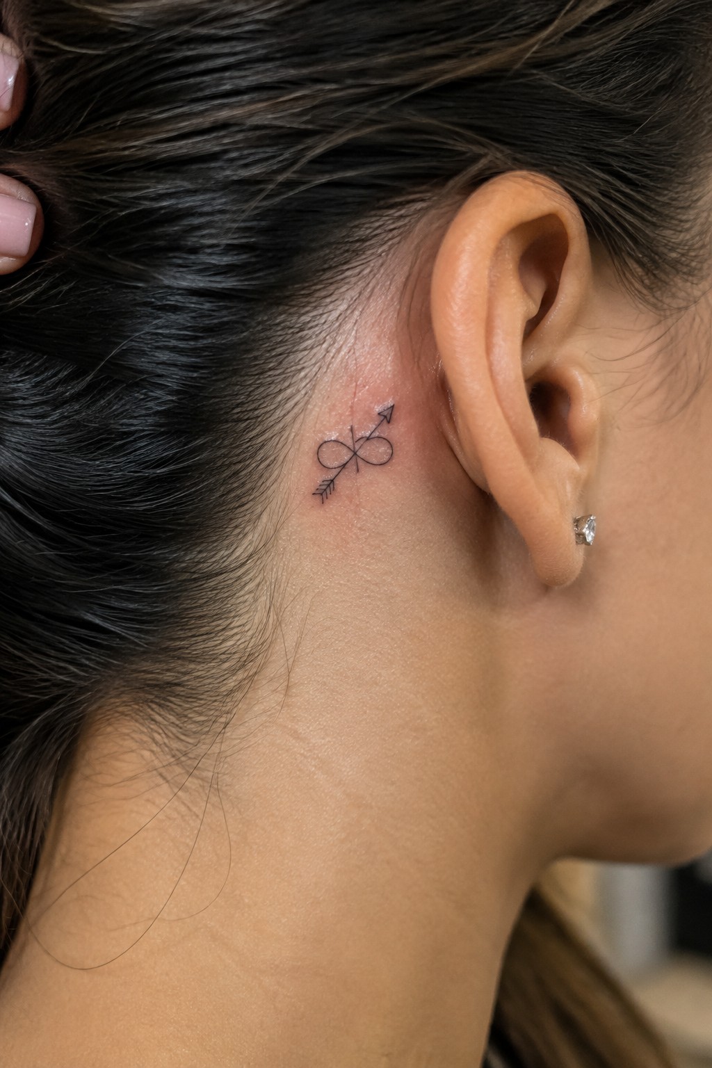Small black infinity tattoo crossed by an arrow behind a woman’s ear.