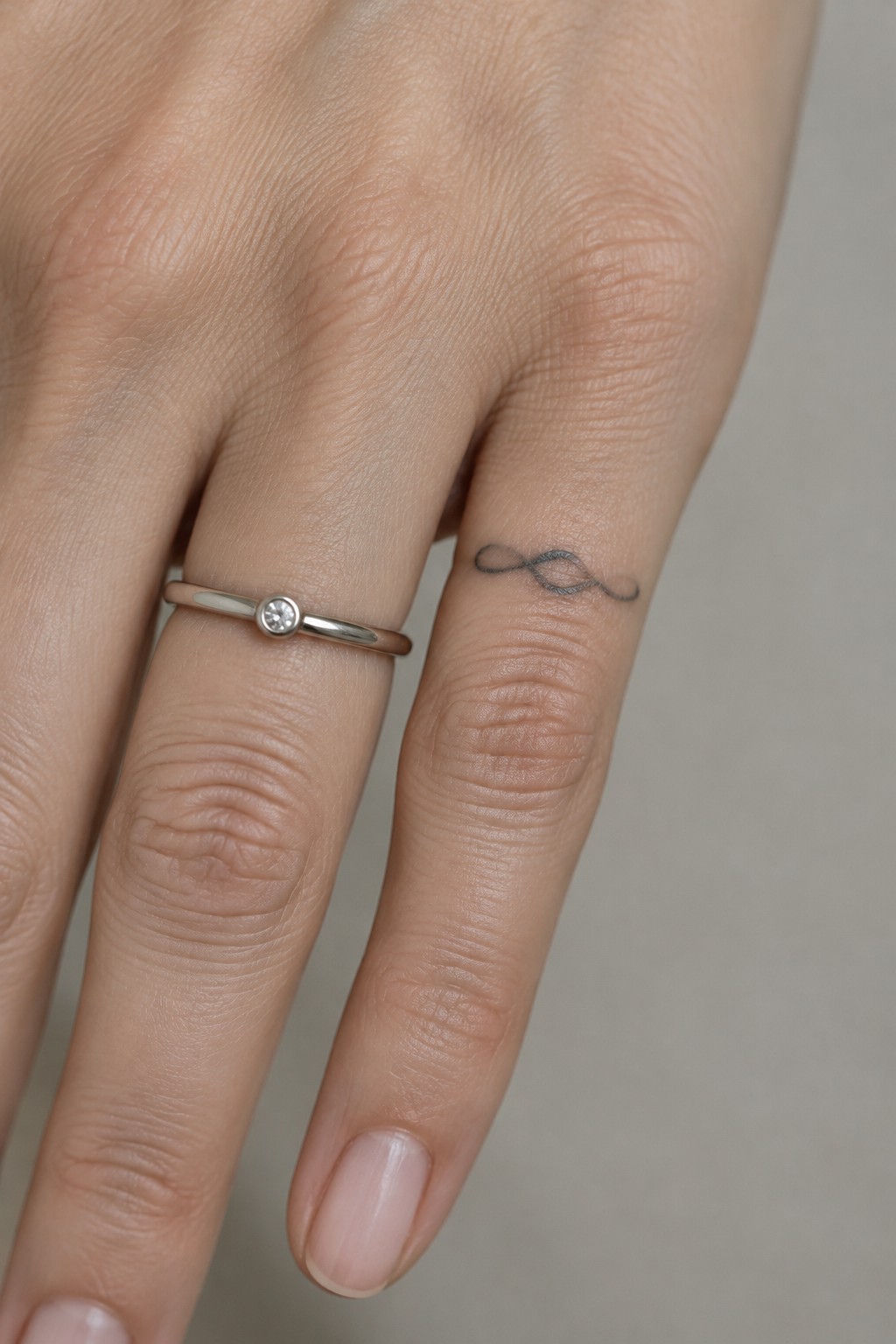 Small black infinity tattoo on the side of a finger beside a thin ring with a round stone.
