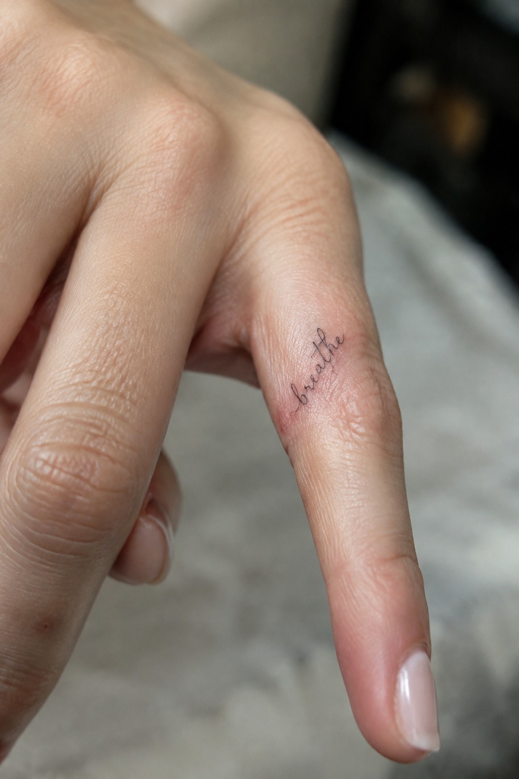 Small fine-line cursive “breathe” tattoo on the side of a finger.