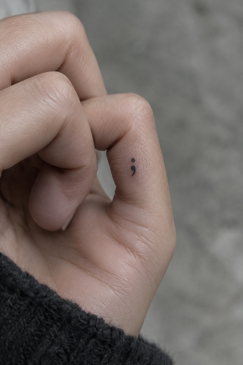 Small black semicolon tattoo on the side of a finger.