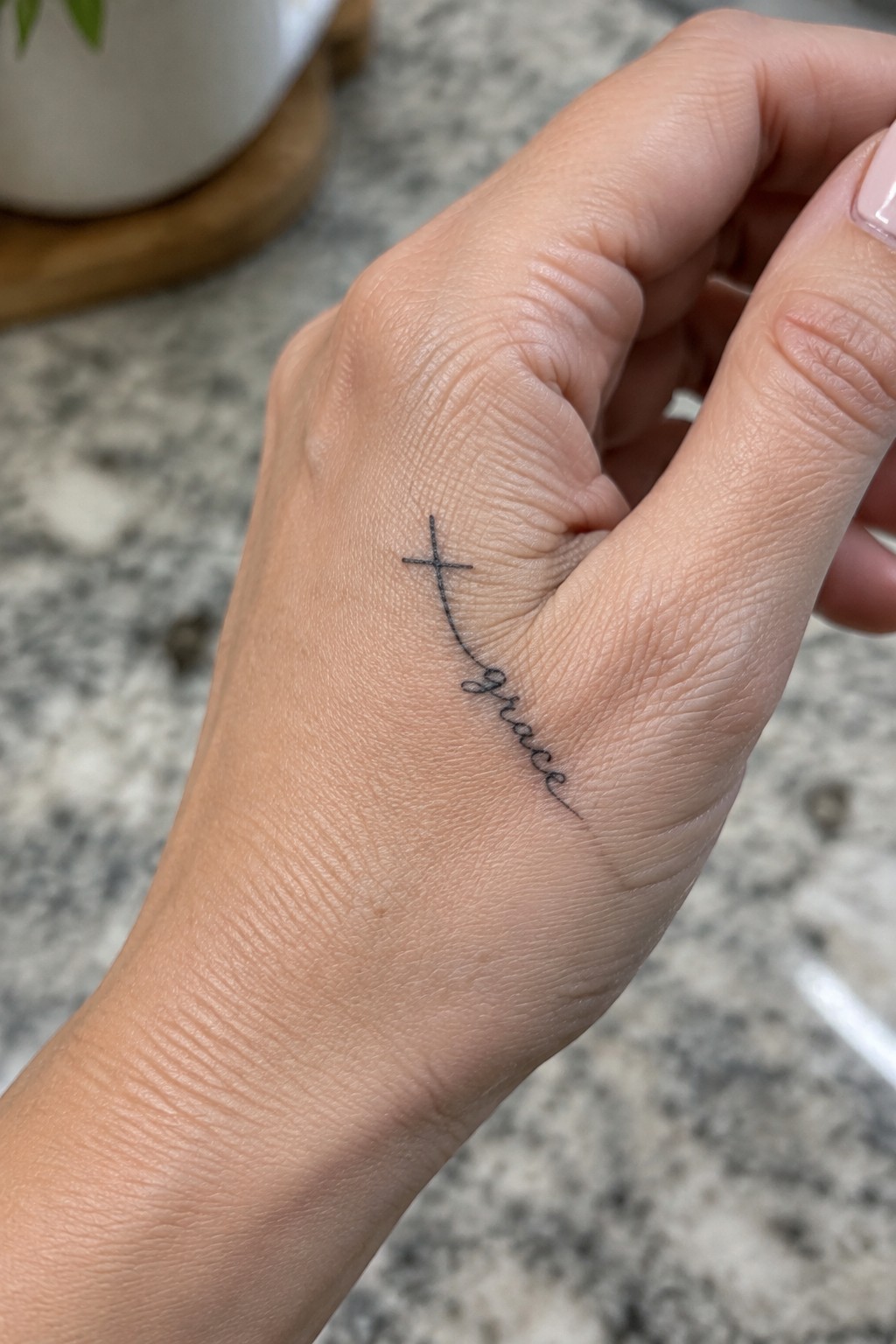 Small fine-line cross connected to cursive “grace” lettering on the side of a hand near the thumb.