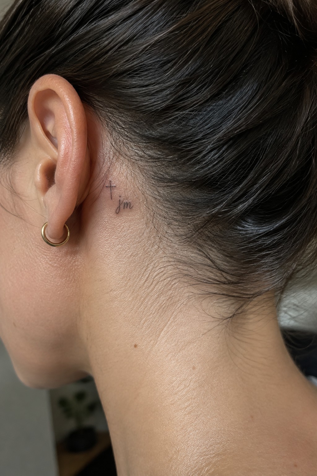 Small fine-line cross and cursive “I’m” tattoo behind a woman’s ear, above a gold hoop earring.