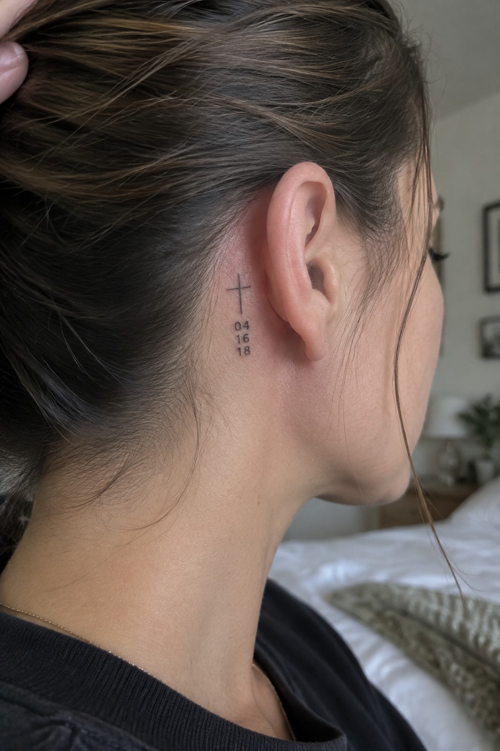 Small fine-line cross tattoo with the date 04 16 18 behind a woman’s ear.