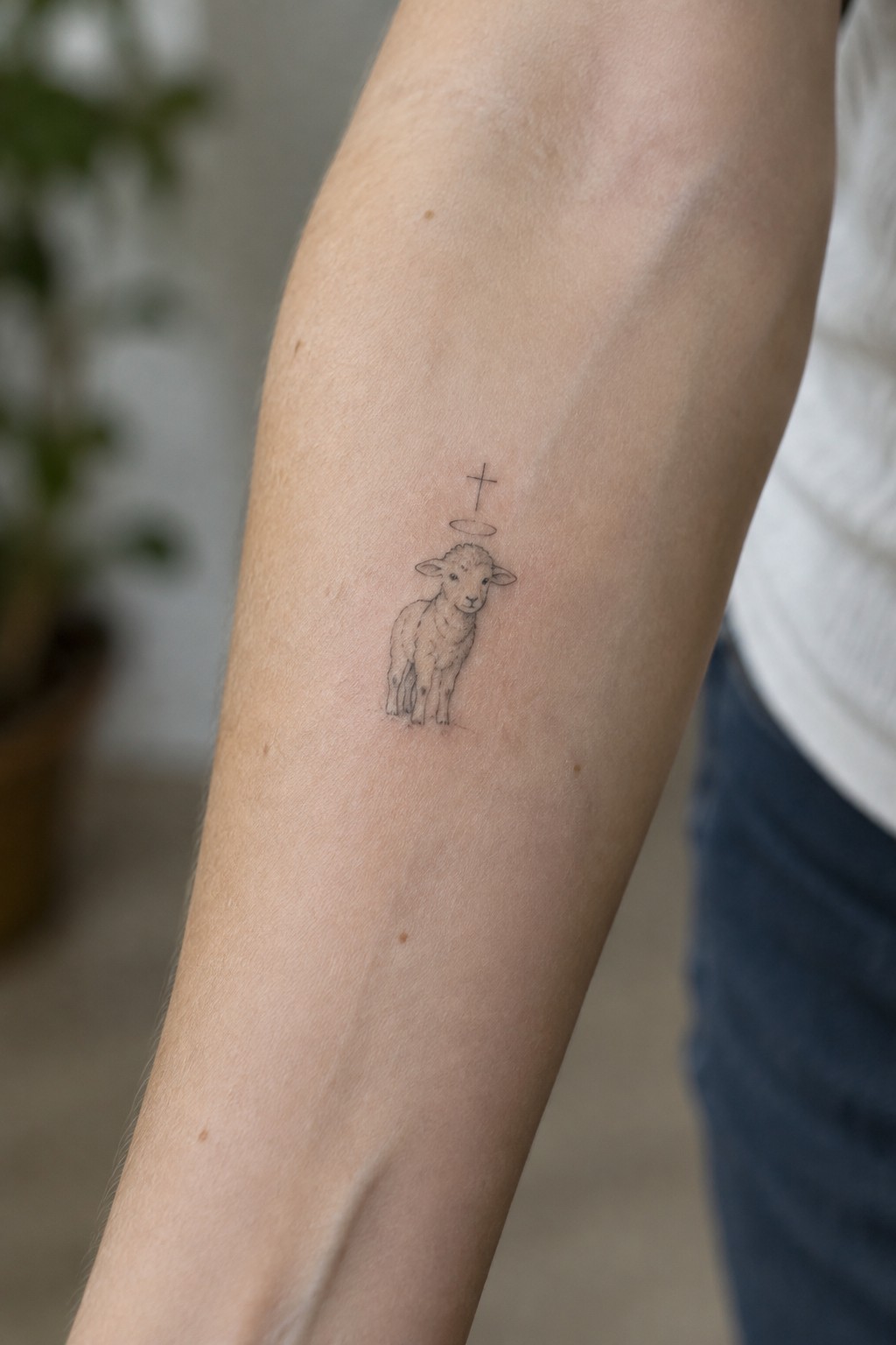 Tiny lamb with halo and cross tattoo on upper arm in white shirt and jeans