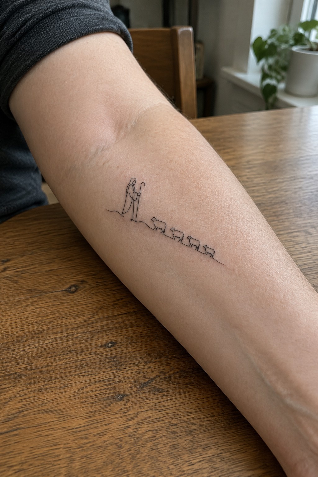 Gray-sleeved forearm with minimalist line tattoo of shepherd and sheep on wooden table