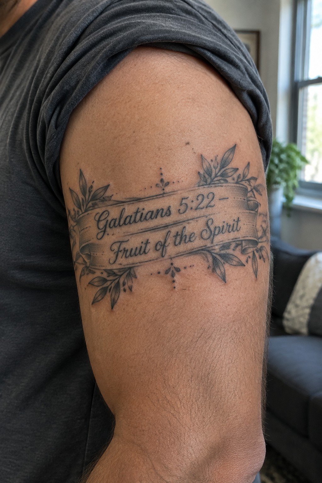 Upper arm tattoo on gray shirt reading Galatians 5:22 Fruit of the Spirit with leaves