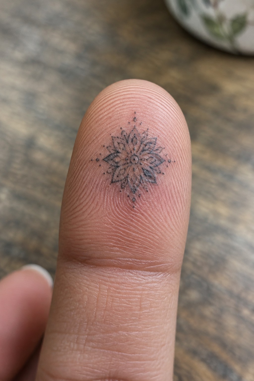 Small black fine-line mandala tattoo with pointed petals and dotted details on the top of a finger.