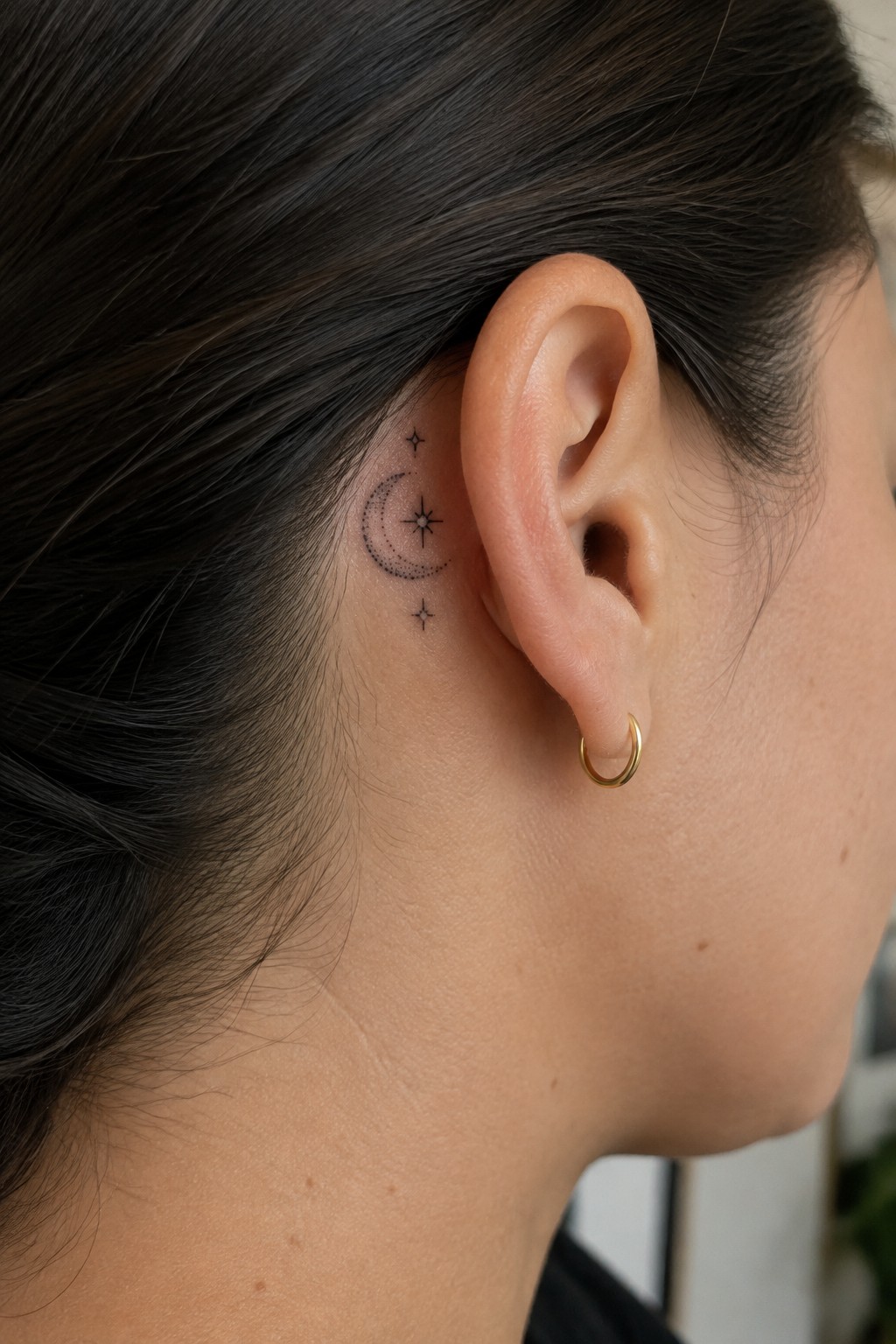 Small black crescent moon tattoo with dotted details and tiny stars behind a woman’s ear, beside a gold hoop earring.