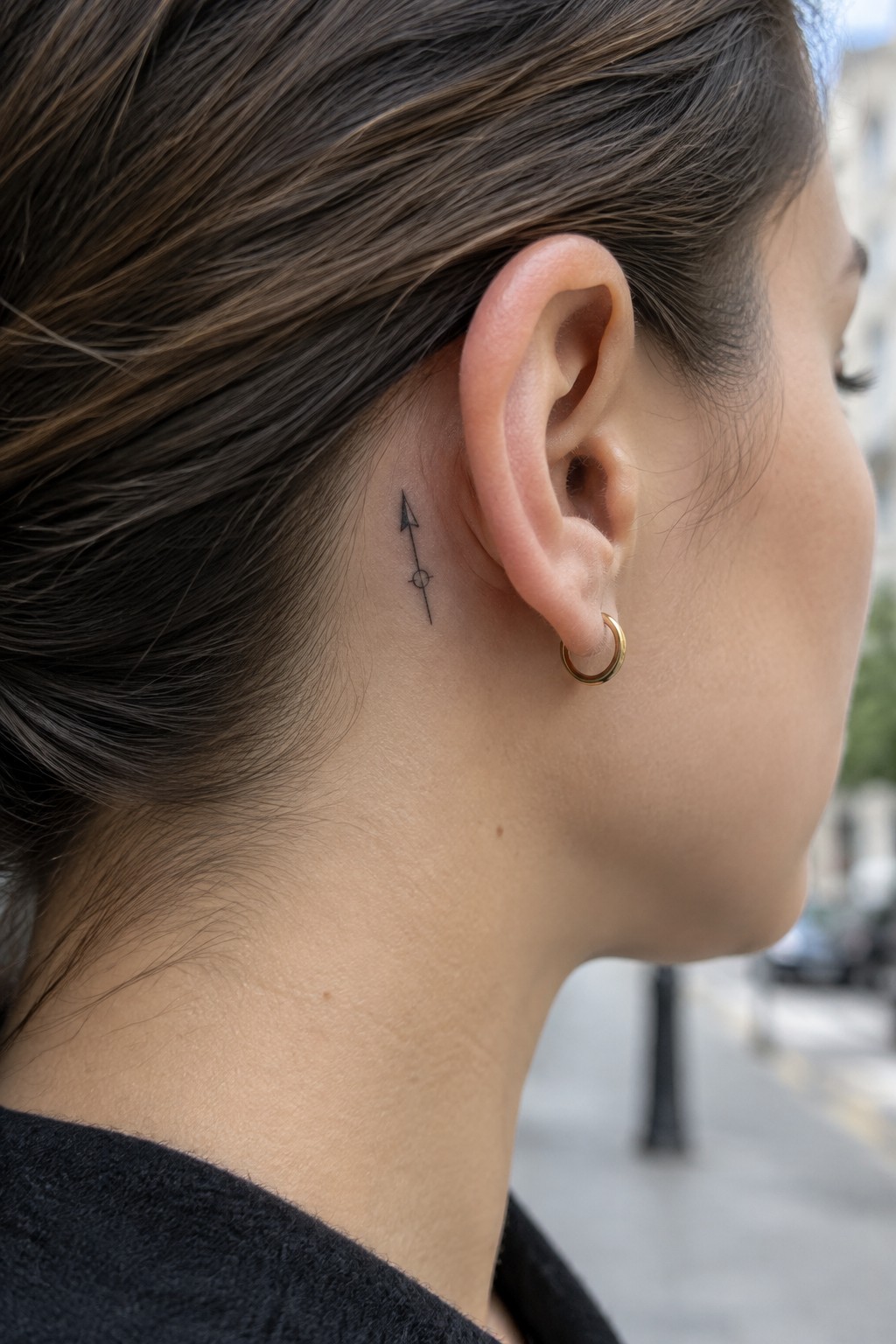 Small fine-line arrow tattoo with a circle on its shaft behind a woman’s ear.