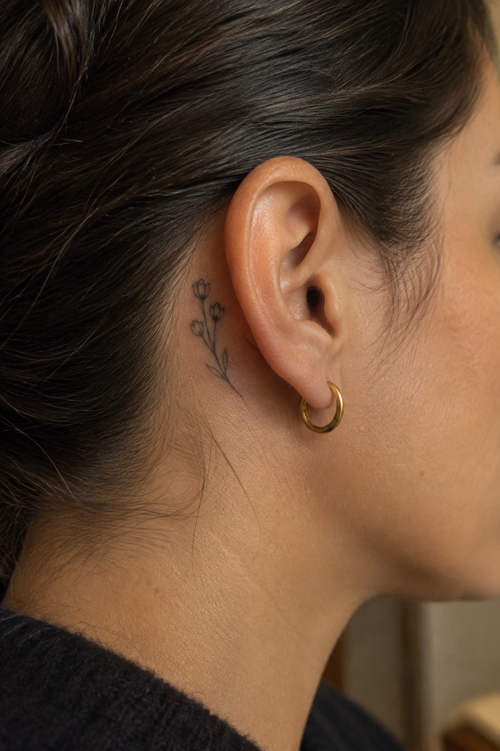 Fine-line tattoo of a small three-bud flower sprig behind a woman’s ear, beside a gold hoop earring.