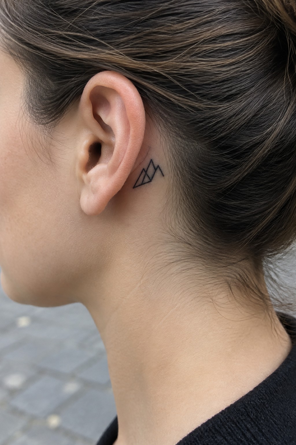 Small black geometric mountain outline tattoo behind a woman’s ear.