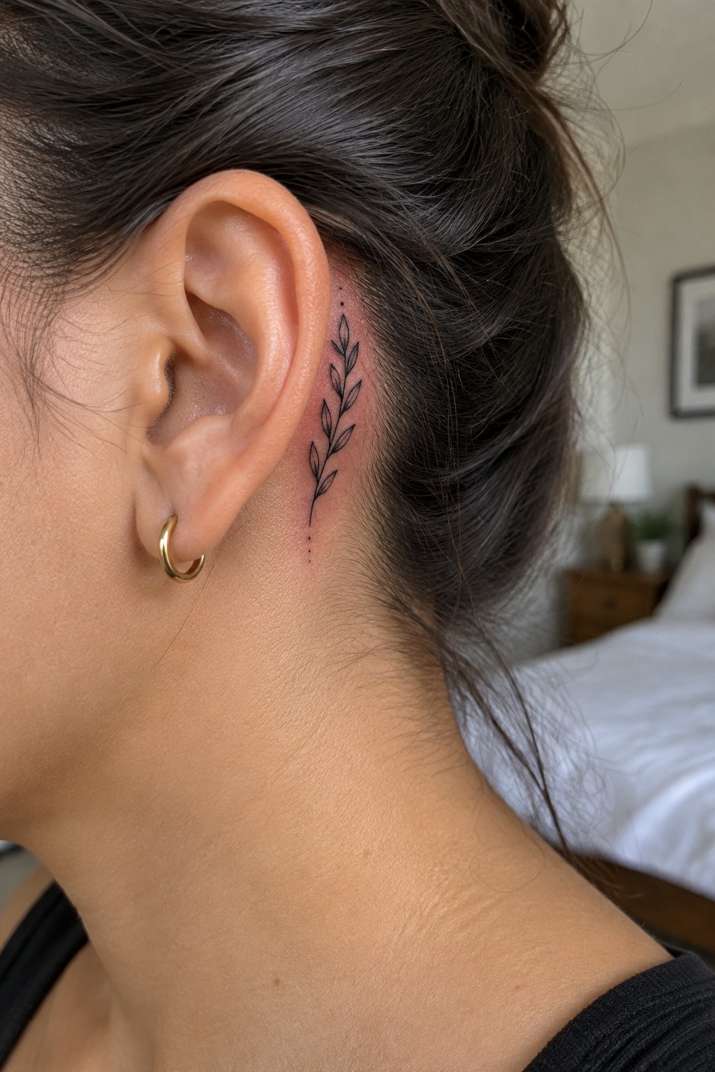 Fine-line leafy sprig tattoo behind a woman’s ear, with a small gold hoop earring.