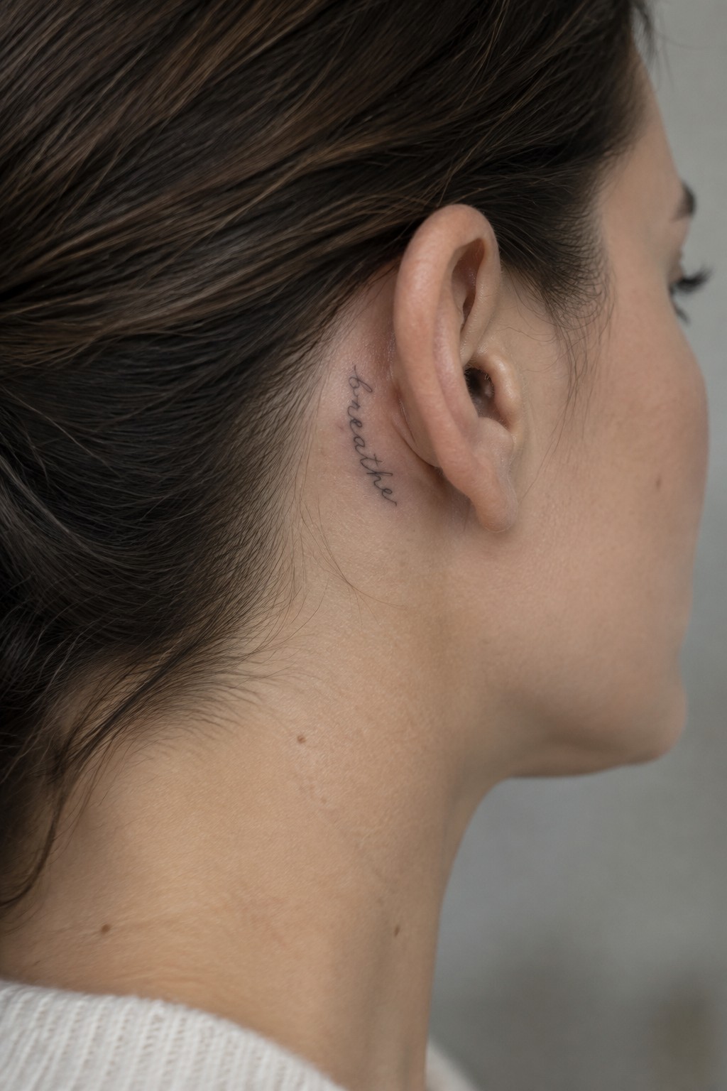 Small fine-line script tattoo reading “breathe” behind a woman’s ear.