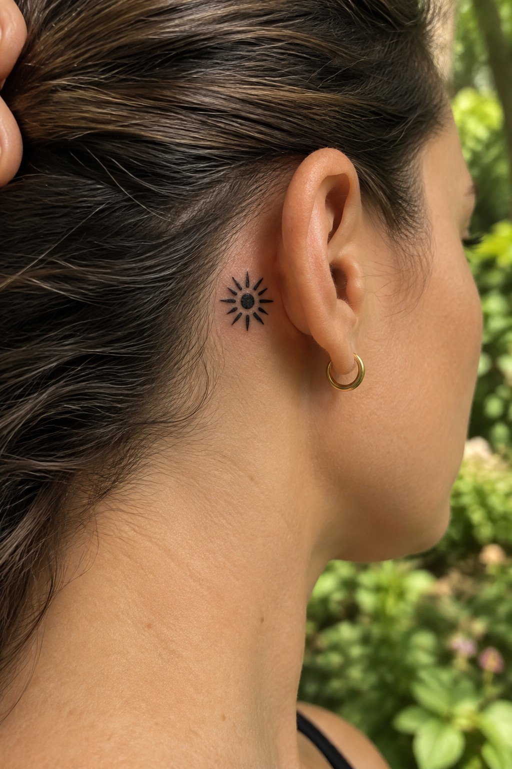 Small black sun tattoo with a solid center and pointed rays behind a woman’s ear.