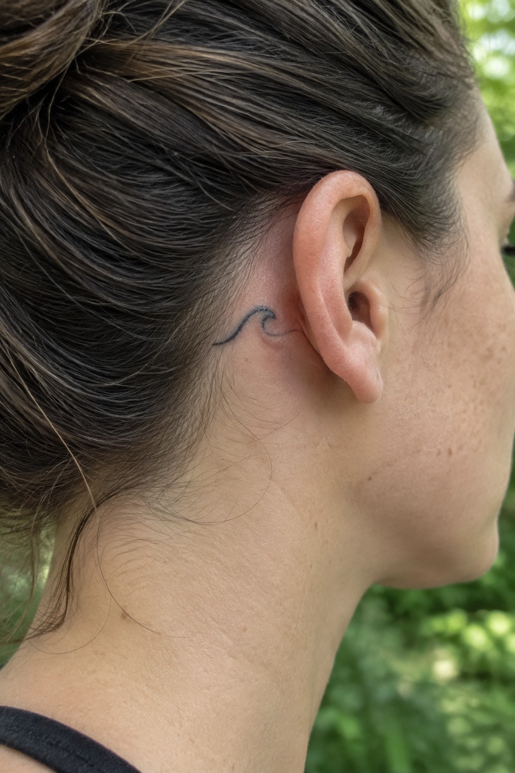 Small black wave tattoo behind a woman’s ear.