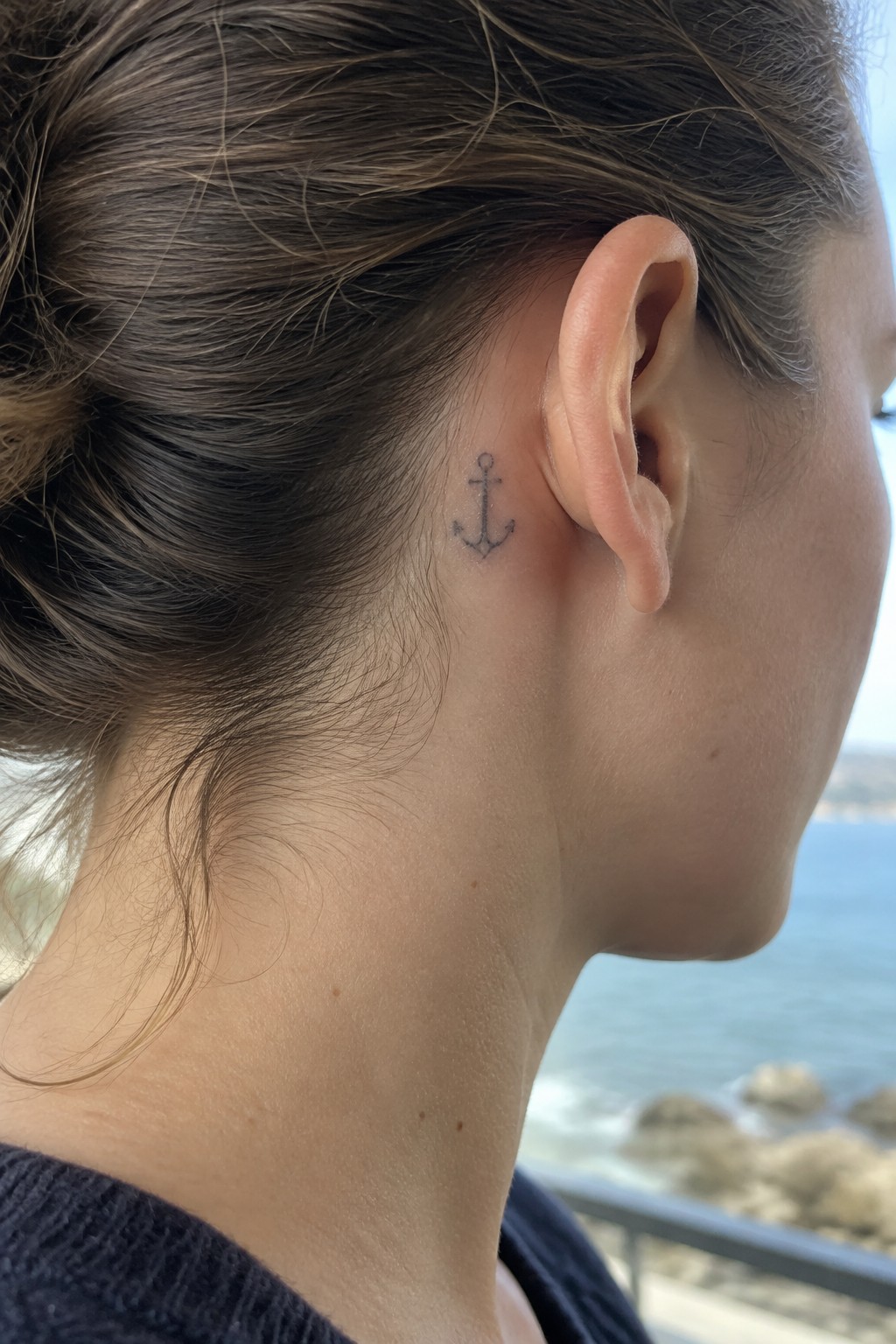 Small fine-line anchor tattoo behind a woman's ear.