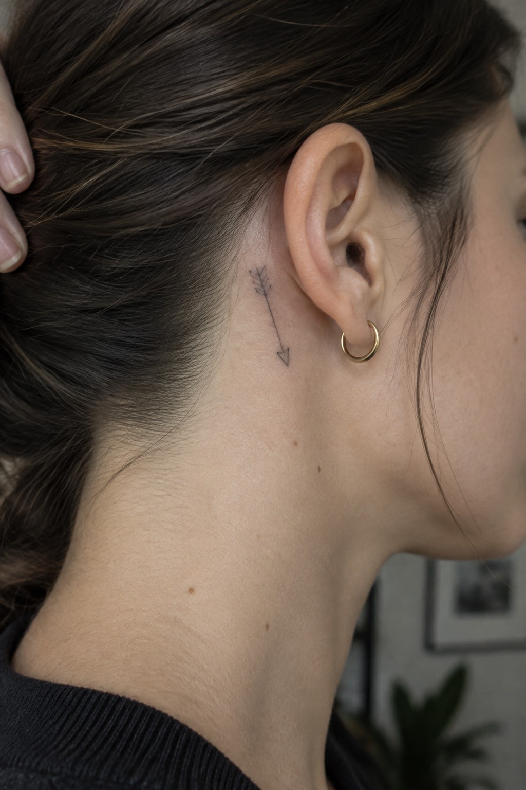 Fine-line arrow tattoo with small feather details behind a woman’s ear, pointing down toward her neck.