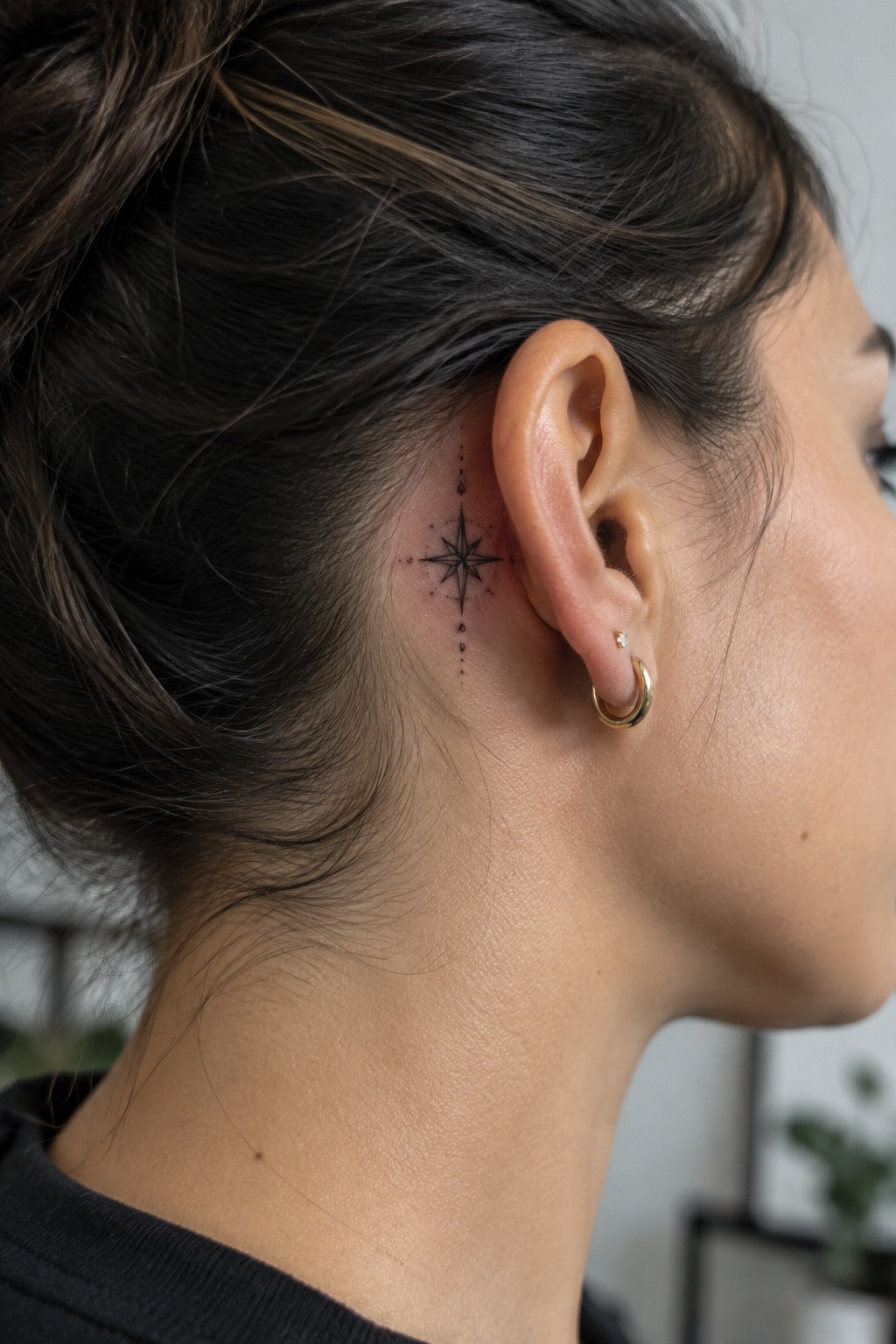 Small black fine-line compass tattoo with dotted accents behind a woman’s ear.