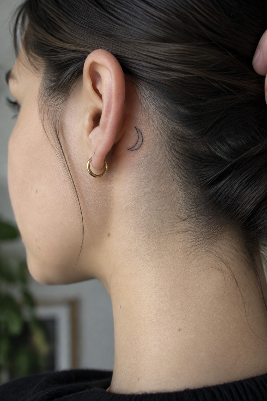 Fine-line crescent moon tattoo behind a woman’s ear, above a small gold hoop earring.