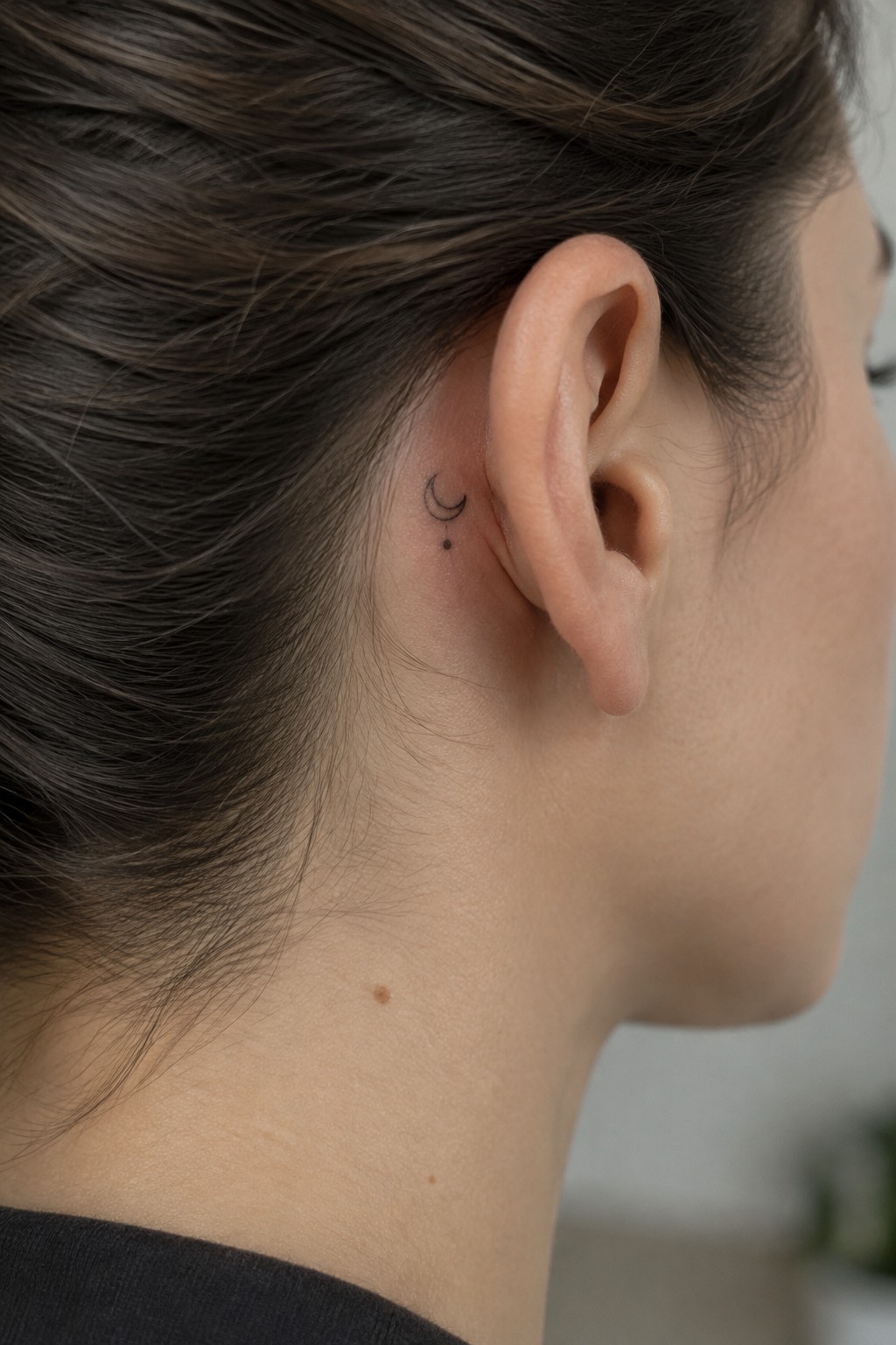 Fine-line crescent moon tattoo with a small dot behind a woman’s ear.