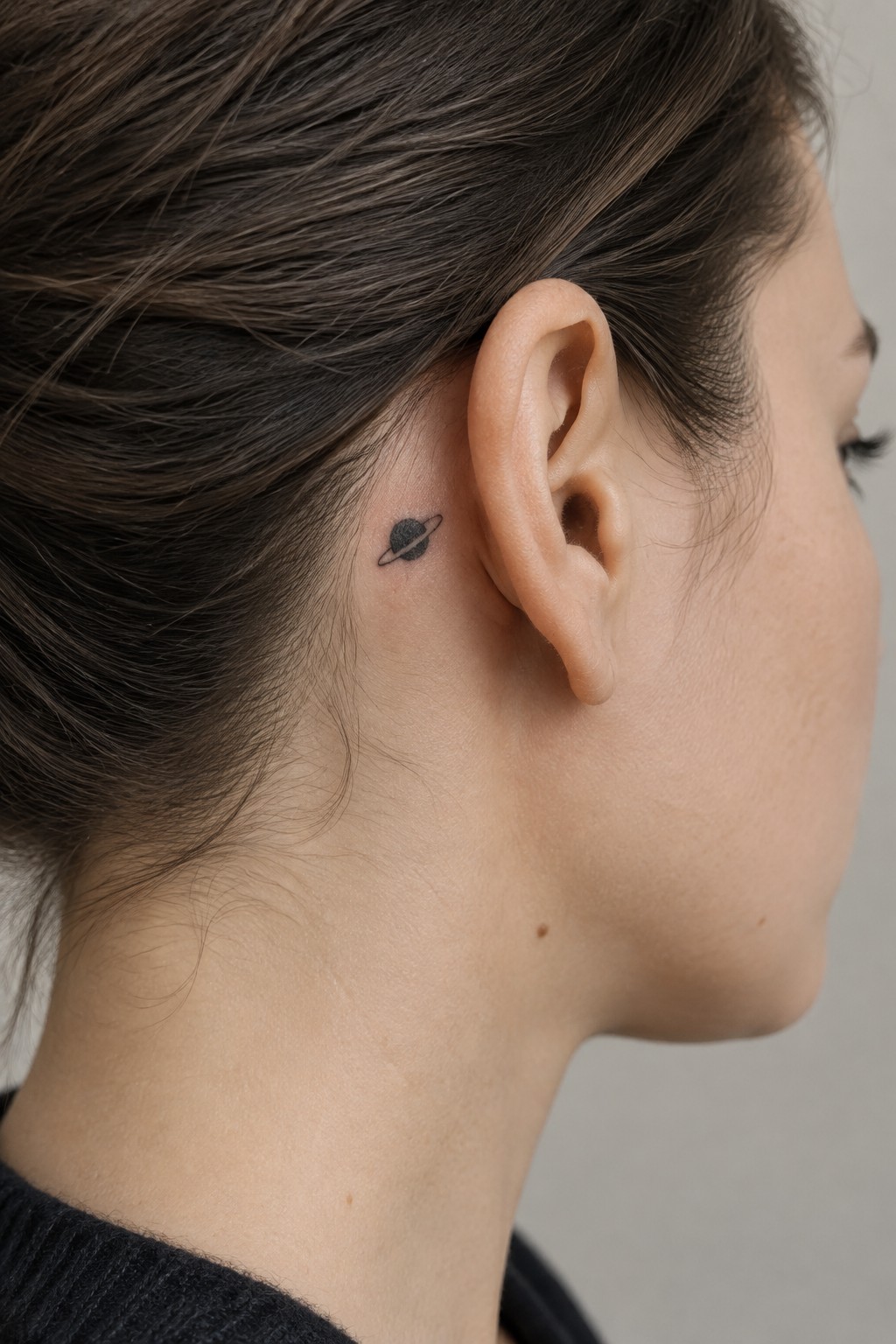 Small black Saturn tattoo behind a woman’s ear, featuring a shaded planet and thin ring.