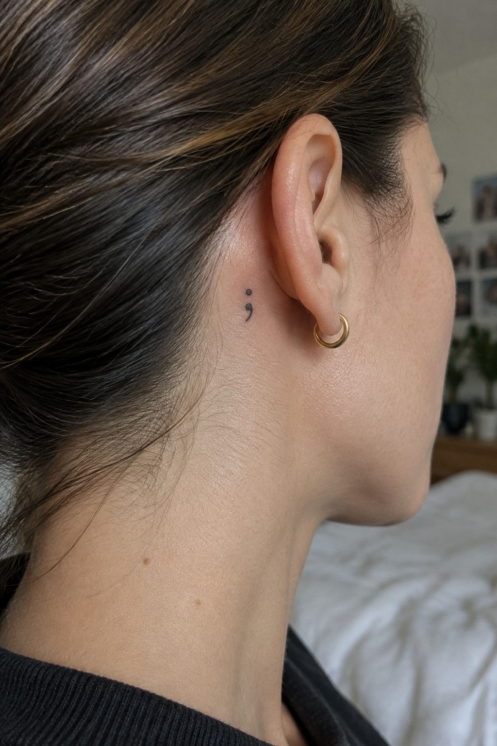 Small black semicolon tattoo behind a woman’s ear, above a gold hoop earring.