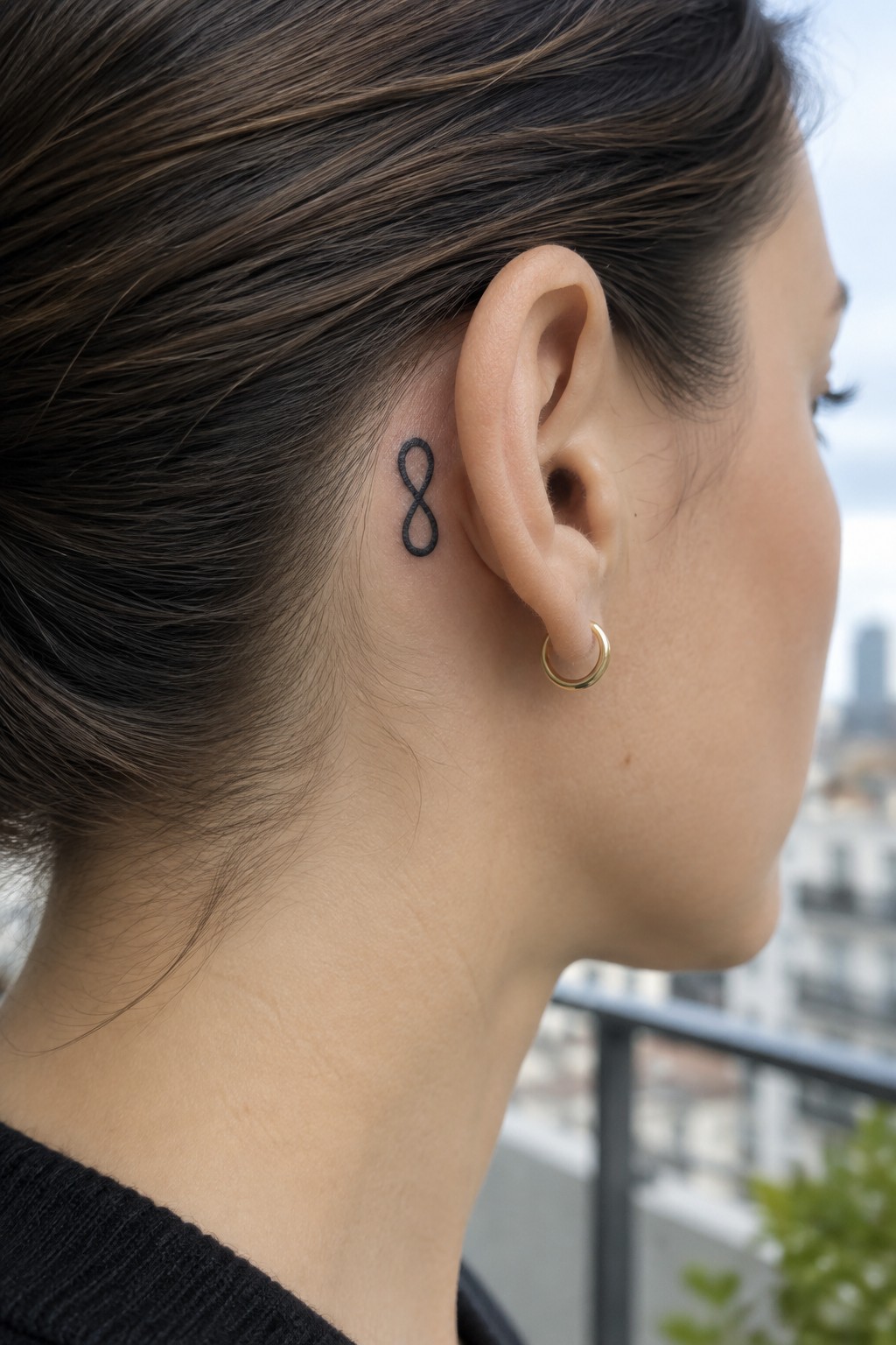 Small black infinity symbol tattoo behind a woman’s ear, above a gold hoop earring.