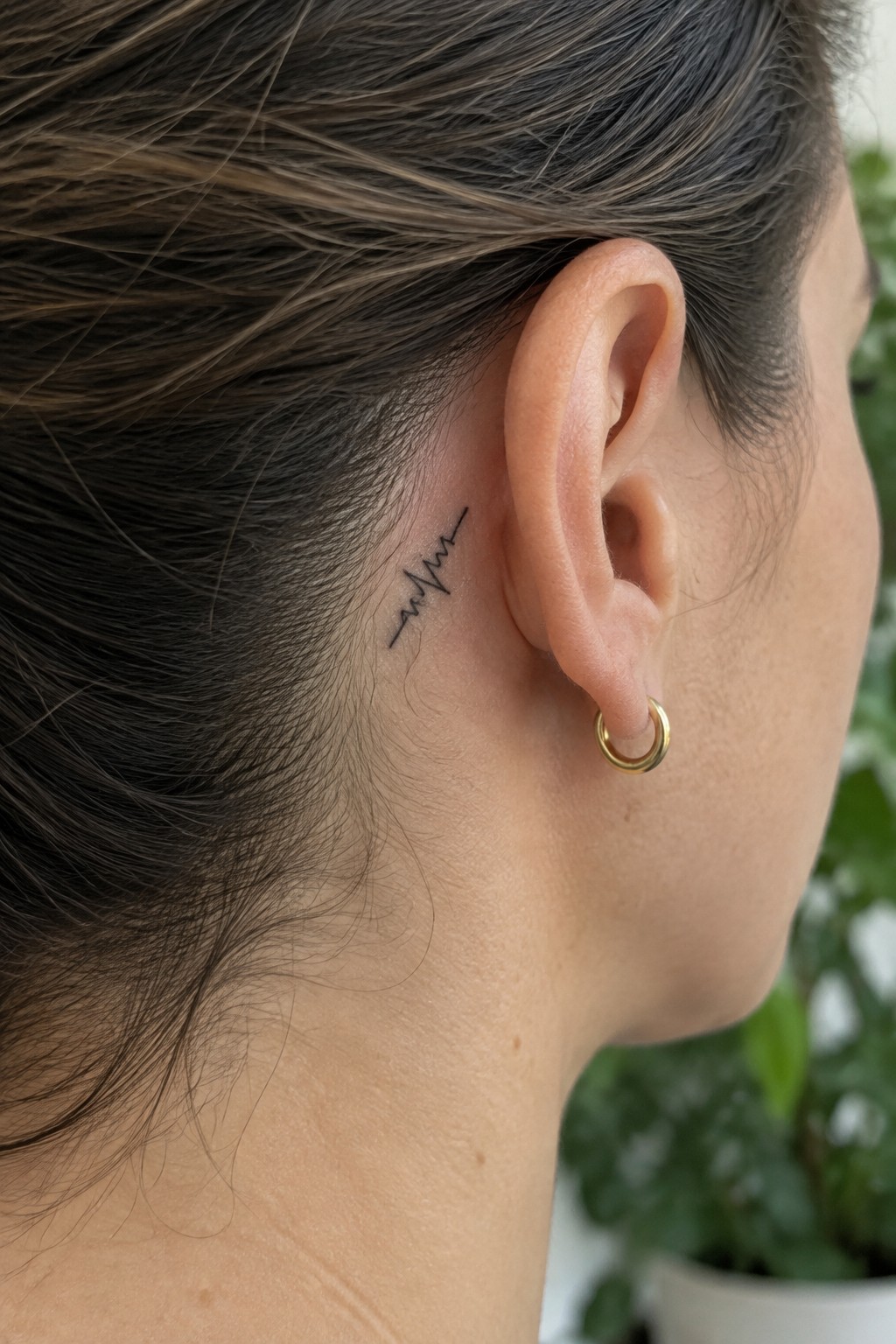Fine black heartbeat line tattoo behind a woman’s ear, above a small gold hoop earring.