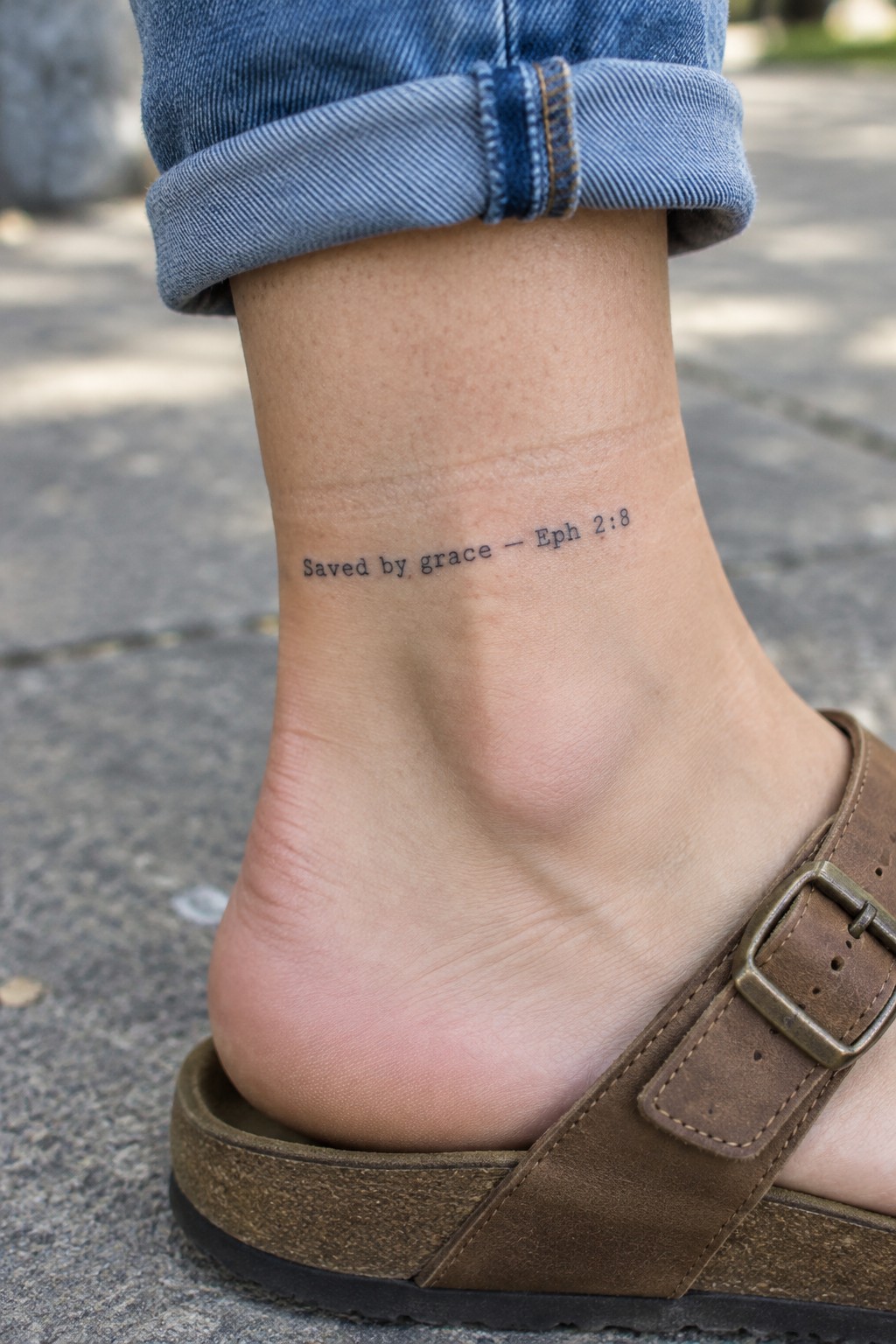 Small black script tattoo on the ankle showing the words saved by grace and the reference Eph 2:8 above a brown sandal.