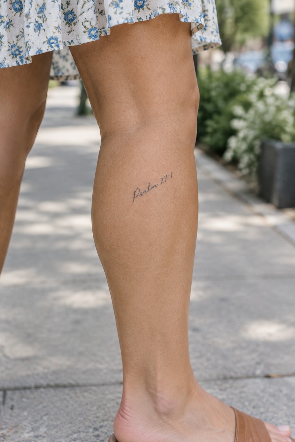 Leg with 'Psalm 27:1' tattoo under floral blue dress and brown sandal