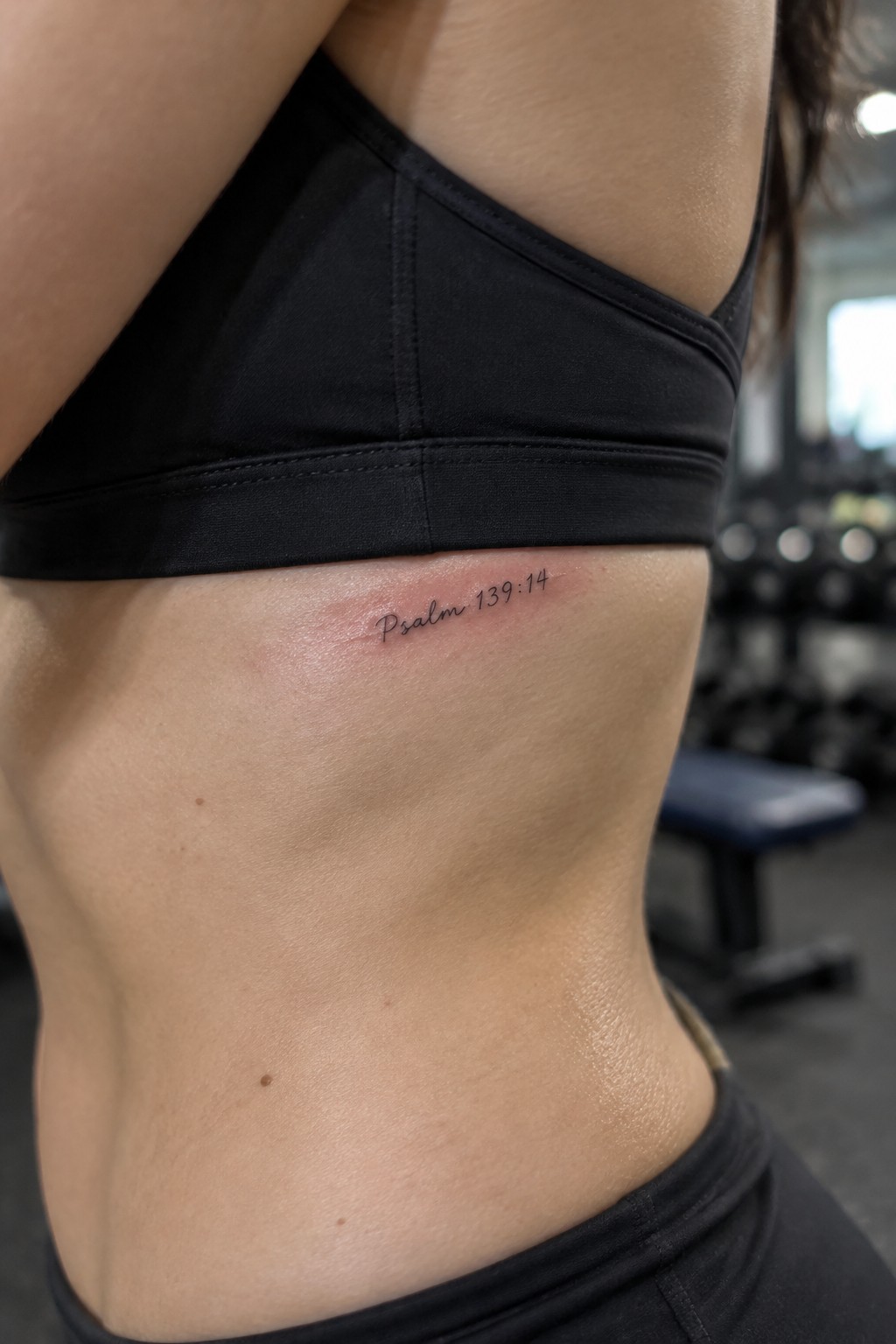 Woman's side with Psalm 139:14 tattoo below black sports bra in gym.