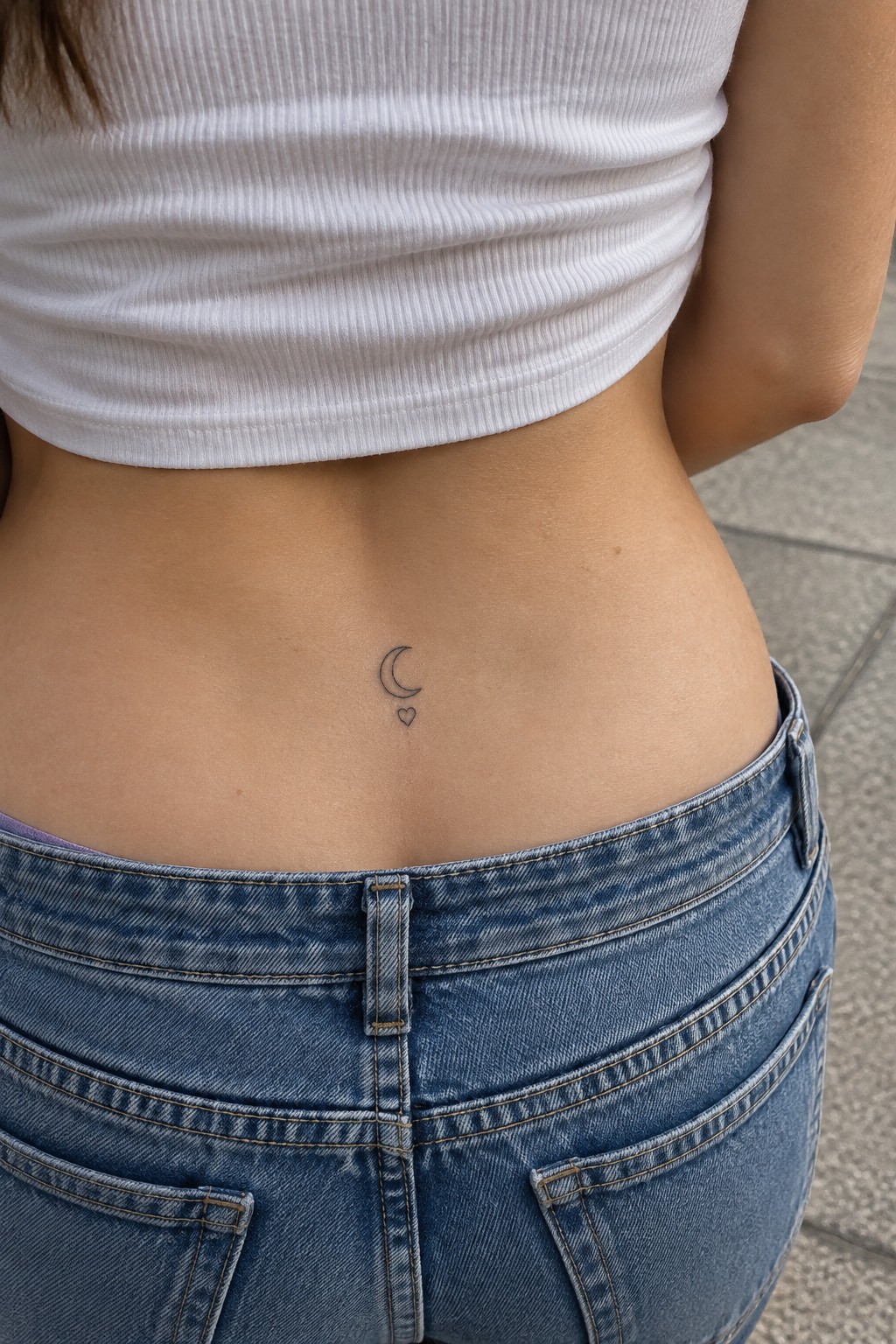 Small fine-line crescent moon and heart tattoo centered on the lower back above blue jeans.