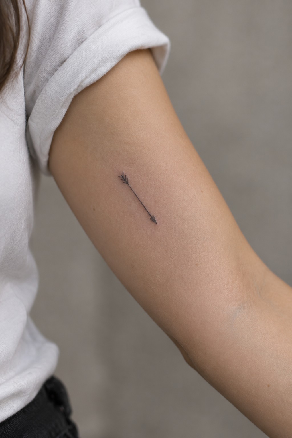 Small black fine-line arrow tattoo on a woman’s upper arm.