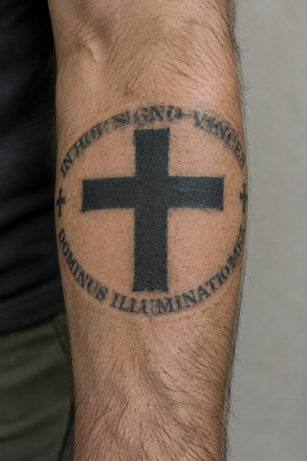 Forearm tattoo with a bold black cross and Latin text arranged in a circle around it