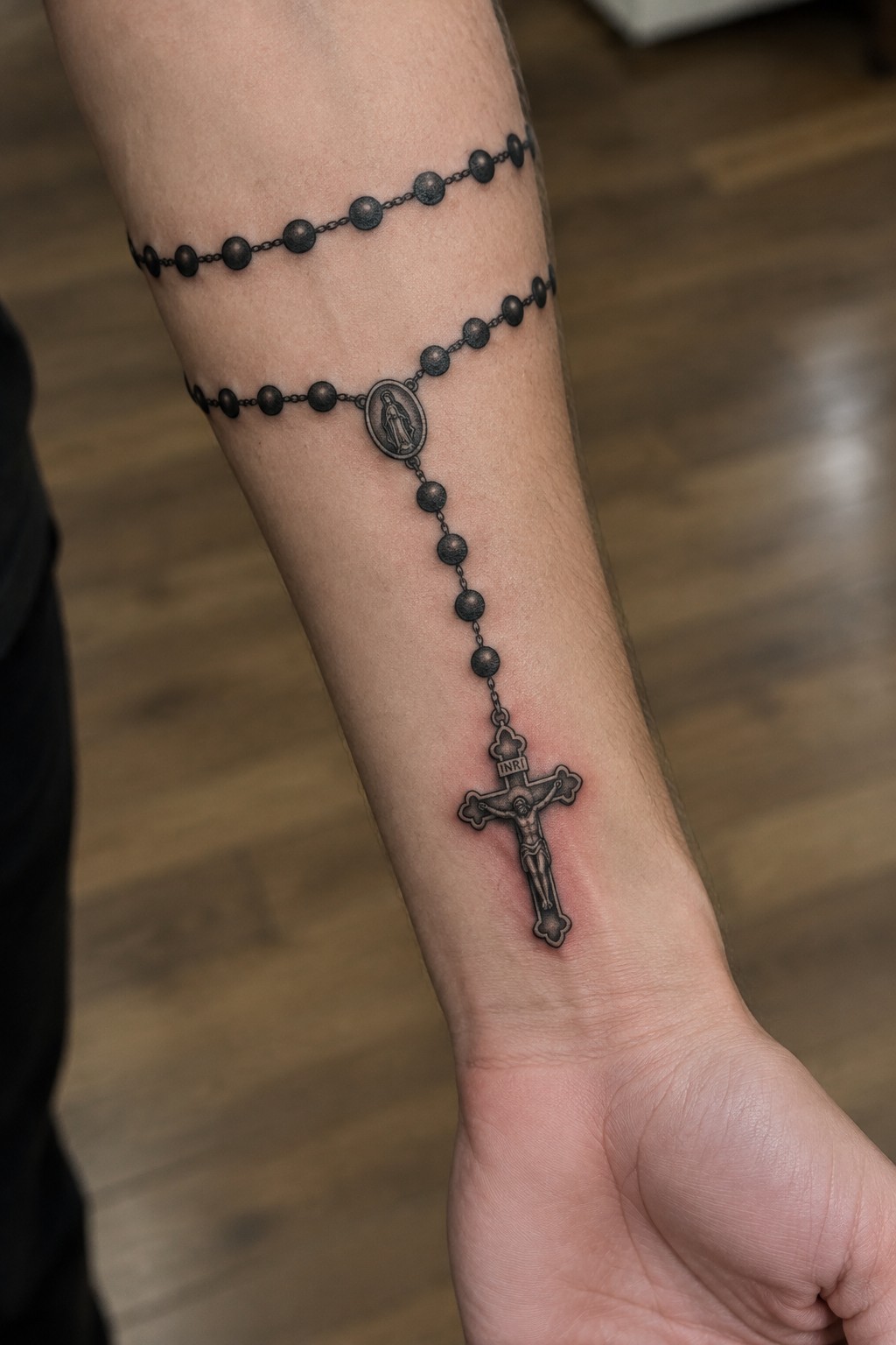 Black and gray rosary tattoo with crucifix and medallion on forearm over wood floor.