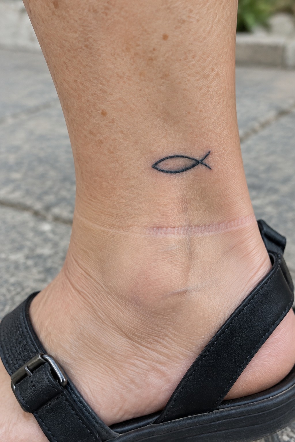 Ankle with small fish tattoo above a thin scar and black sandal strap on pavement