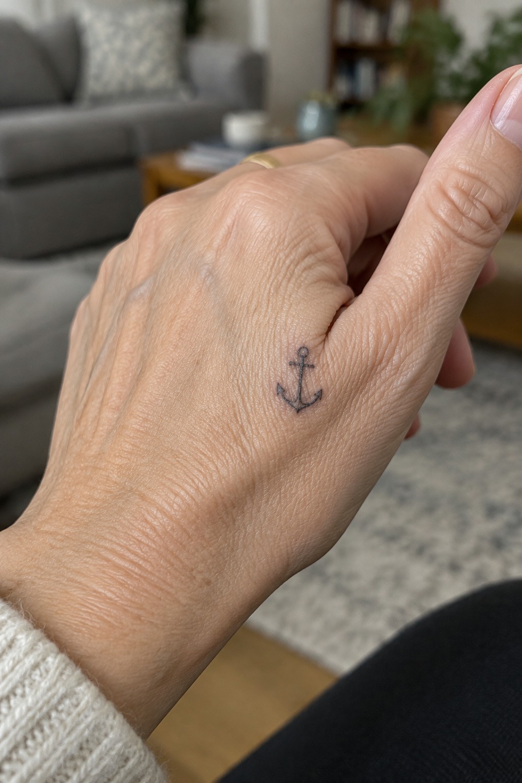 Hand with small black anchor tattoo, gold ring, and white knit sleeve