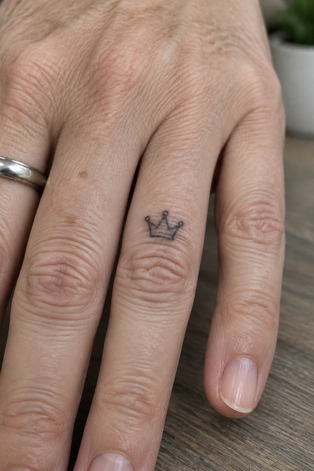 Small black crown tattoo on the side of a ring finger next to a silver band.