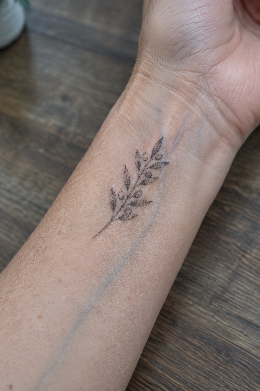 Black ink leafy branch tattoo with berries on light-skinned forearm over wood