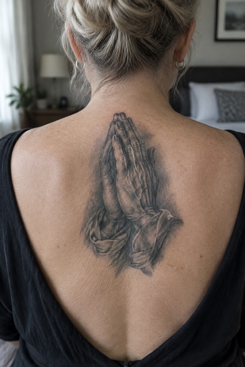 Realistic black and grey tattoo of praying hands with draped fabric centered on a woman's upper back.