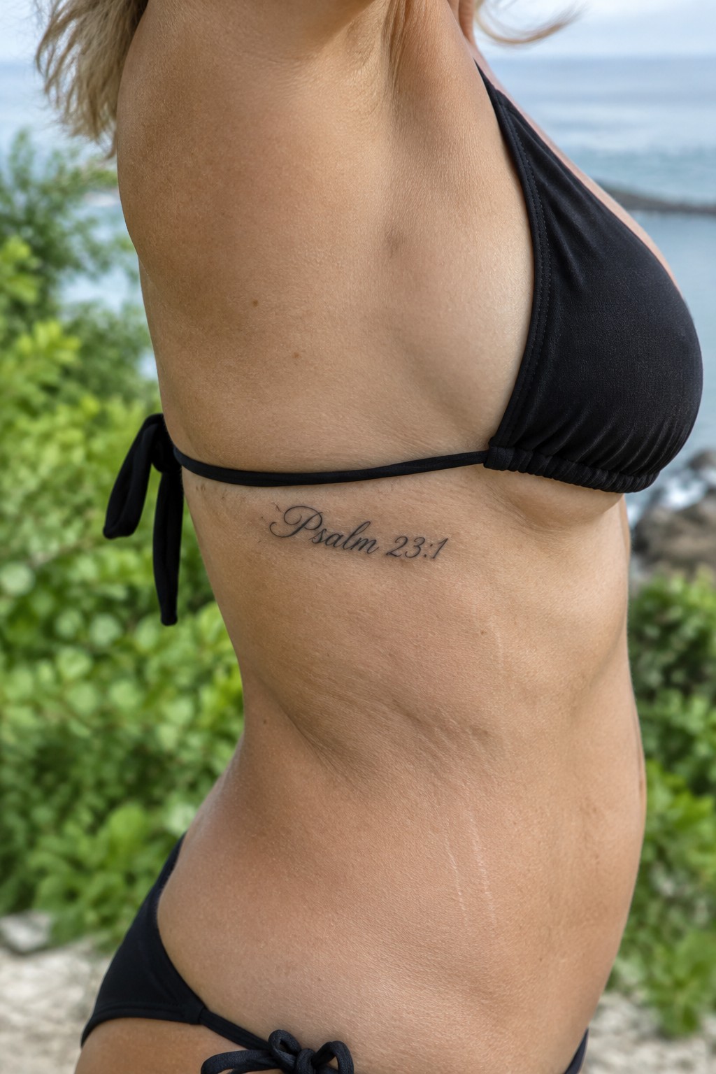Woman in black bikini with Psalm 23:1 tattoo on side by ocean