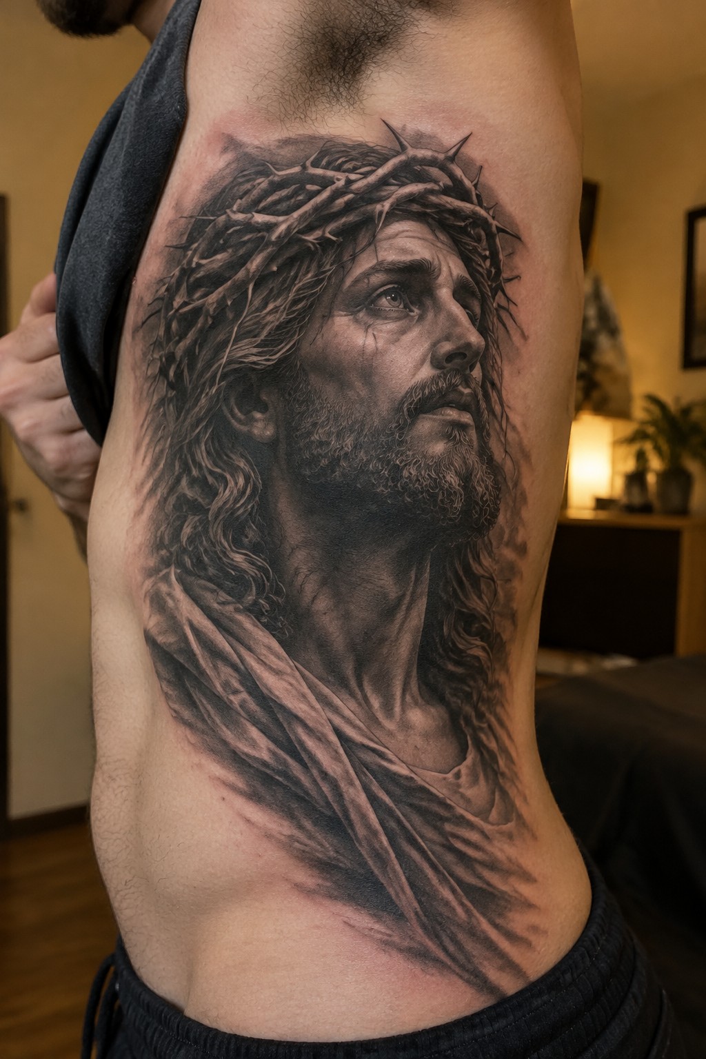 Black and grey realistic tattoo of Jesus with crown of thorns on a man's side torso, showing the full portrait from chest to waist