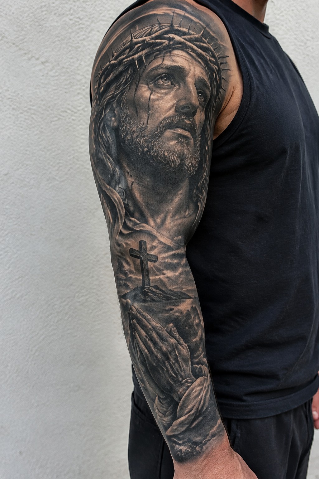 Realistic black and gray tattoo of Jesus with crown of thorns on a man's upper arm, extending into praying hands and a cross on the forearm