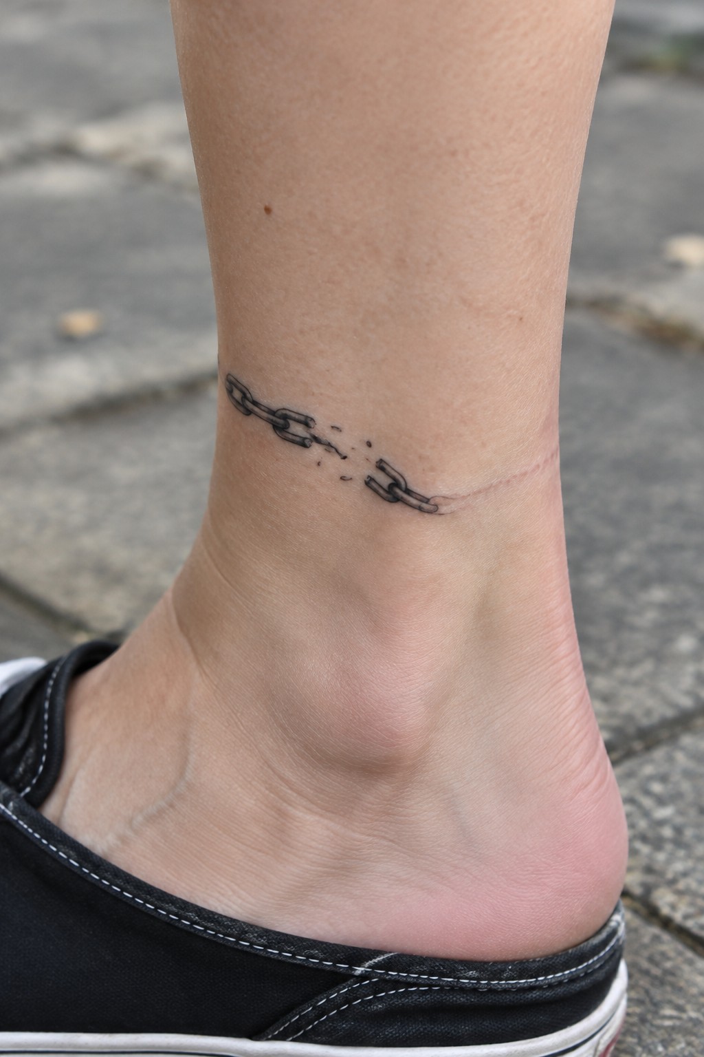 Small black broken chain tattoo curving around the ankle above a black shoe.