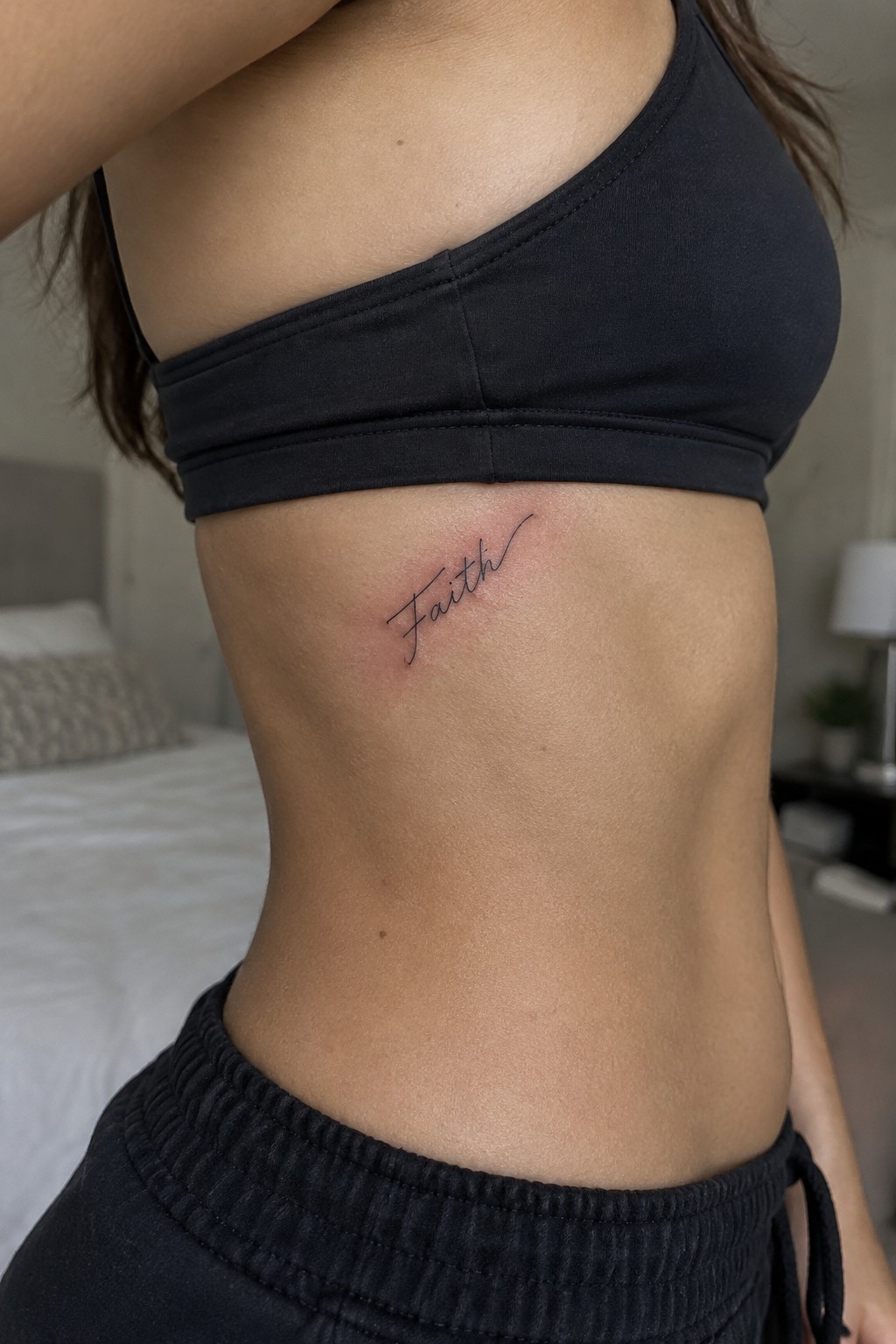 Small black script tattoo reading “Faith” on the side of a woman’s ribs beneath a black bra.
